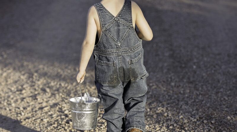Bambino biondo che cammina lungo un sentiero con secchiello e canna da pesca. Di spalle.